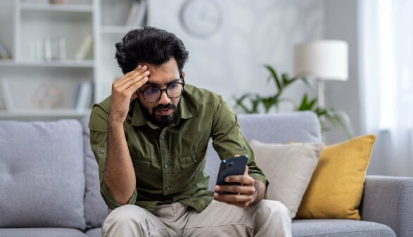 Ein Mann sitzt auf dem Sofa und schaut verzweifelt auf sein Smartphone.