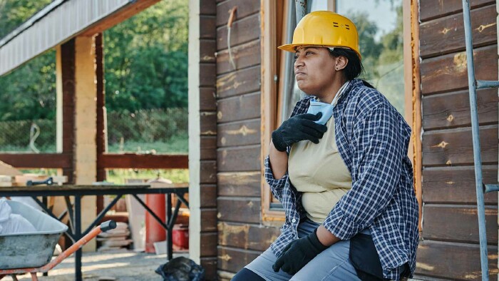 Eine Handwerkerin in Arbeitshose, kariertem Hemd und mit Bauhelm sitzt am Rand einer Baustelle. Sie blickt angestrengd und erschöpft in die Ferne.