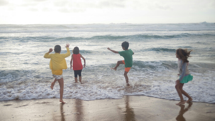 Vier Kinder spielen ausgelassen am Strand im Bereich der Brandung. Sie stehen mit dem R&uuml;cken zur Kamera und tragen bunte Sommerkleider in Gelb, Rot, Gr&uuml;n und Hellblau. Die Wellen rollen auf sie zu, eines der Kinder springt &uuml;ber das Wasser. Der Himmel ist leicht bew&ouml;lkt, das Meer rau.