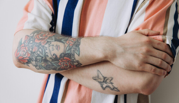 Nahaufnahme verschr&auml;nkter Arme einer Person im gestreiften Hemd. Der linke Unterarm ist gro&szlig;fl&auml;chig t&auml;towiert mit Rosen, einem Frauengesicht und Schriftz&uuml;gen im traditionellen Tattoo-Stil. Auf dem rechten Unterarm ist ein kleineres Stern-Tattoo zu sehen.