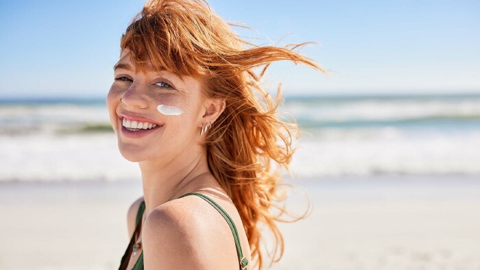 Junge Frau mit Sommersprossen und Sonnencreme im Gesicht lächelt am Strand bei Sonnenschein – Sonnenschutz, Sonnencreme-Test, Hautpflege im Sommer.