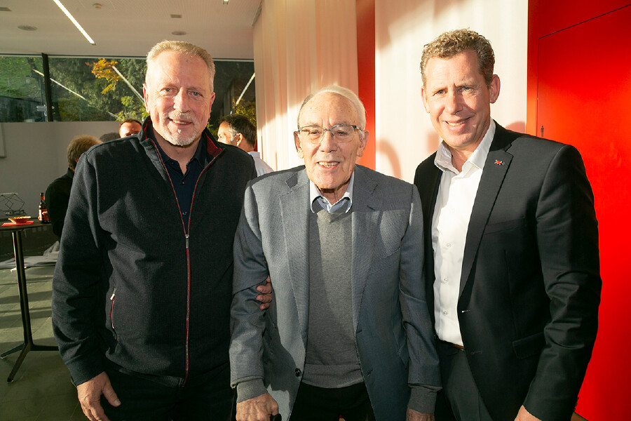 Drei M&auml;nner stehen nebeneinander in einem hellen Raum und l&auml;cheln in die Kamera. Links ein Mann in einer dunklen Zipper-Jacke, in der Mitte ein &auml;lterer Mann mit Brille, grauem Sakko und Pullover (Bertram J&auml;ger), und rechts ein Mann in einem dunklen Anzug mit wei&szlig;em Hemd (Bernhard Heinzle). Im Hintergrund ist eine markante rote Wand und eine Fensterfront zu sehen.