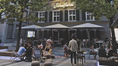 Abendliche Au&szlig;enansicht des Geb&auml;udes der Schaffarei im AK Quartier. Im geschm&uuml;ckten Au&szlig;enbereich sitzen zahlreiche Personen an Tischen unter gro&szlig;en wei&szlig;en Schirmen sowie auf Liegest&uuml;hlen. &Uuml;ber der Fassade leuchtet der Schriftzug &bdquo;Schaffarei&ldquo; in gelber Neonschrift.