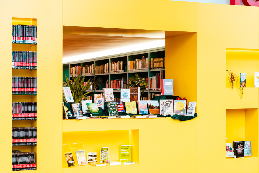 Innenansicht der AK Bibliothek: Eine gro&szlig;e, leuchtend gelbe Wand mit mehreren Nischen dient als Pr&auml;sentationsfl&auml;che. In einer zentralen &Ouml;ffnung sind zahlreiche B&uuml;cher frontal aufgestellt und ausgestellt. Durch den Durchblick dahinter sind gr&uuml;ne Regalreihen voller B&uuml;cher sichtbar. In schmalen seitlichen Wandnischen stehen weitere Fachb&uuml;cher und Brosch&uuml;ren &uuml;bersichtlich geordnet.