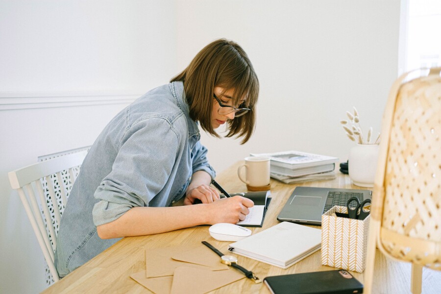 Eine junge Frau sitzt an einem Schreibtisch vor einem Laptop und schreibt in ein Buch.