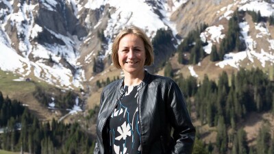 Eine Frau mit halblangen, blonden Haaren, Angela Schwarzmann, lächelt in die Kamera. Im Hintergrund sieht man ein beeindruckendes Bergpanorama, auf dem teils noch Schnee liegt.
