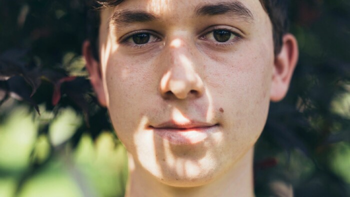 „Porträt eines jungen Gartenbau-Lehrlings mit kurzen braunen Haaren, aufgenommen im natürlichen Licht eines Gartens. Schatten von Blättern fallen über das Gesicht und erzeugen ein lebendiges Spiel aus Licht und Natur. Idealbild für Ausbildung im Garten- und Landschaftsbau, Nachwuchsförderung und grüne Berufe.