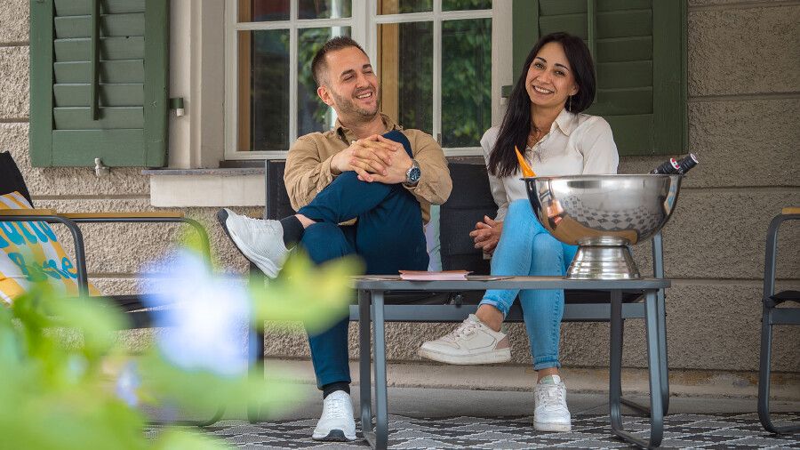 Portr&auml;t von Adrien und Alyssa, den neuen P&auml;chtern der Schaffarei Gastronomie x Aventure im AK Quartier. Beide sitzen entspannt und fr&ouml;hlich auf einer schwarzen Gartenbank im Au&szlig;enbereich vor einem Fenster mit gr&uuml;nen Fensterl&auml;den. Links sitzt Adrien im beigen Hemd, dunkler Hose und wei&szlig;en Sneakern, die Beine &uuml;bereinandergeschlagen. Rechts sitzt Alyssa mit langen, dunklen Haaren, wei&szlig;er Bluse, hellblauen Jeans und ebenfalls wei&szlig;en Sneakern. Auf dem dunklen Tisch vor ihnen steht eine gro&szlig;e, gl&auml;nzende Metallschale zur Flaschenk&uuml;hlung. Im verschwommenen Vordergrund sind gr&uuml;ne Bl&auml;tter zu sehen.