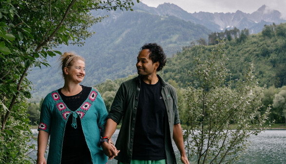 Ein l&auml;chelndes Paar h&auml;lt H&auml;ndchen und blickt sich liebevoll an, w&auml;hrend es in einer alpinen Landschaft spazieren geht. Links steht eine Frau mit hochgesteckten blonden Haaren, einem Haarband und einer markanten t&uuml;rkisfarbenen Strickjacke mit rosa-wei&szlig;en H&auml;kelmustern. Rechts steht ein Mann mit dunklen, lockigen Haaren, einem schwarzen T-Shirt und einem offenen, dunkelgr&uuml;nen Hemd. Im Hintergrund erstreckt sich ein ruhiger See, hinter dem dicht bewaldete Bergh&auml;nge und schroffe, wolkenverhangene Gebirgsz&uuml;ge aufragen. Am linken und rechten Bildrand rahmen gr&uuml;ne Str&auml;ucher und B&auml;ume die Szene ein.