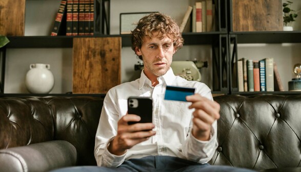 Ein Mann sitzt mit traurigem Blick auf einem Sofa und hält sein Smartphone und seine Kreditkarte in der Hand.