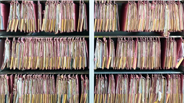 H&auml;ngeregister-Archiv mit zahlreichen prall gef&uuml;llten, rosa Aktenordnern, die eng aneinandergereiht in mehreren Regalf&auml;chern h&auml;ngen.