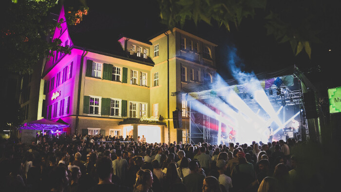 N&auml;chtliches Open-Air-Konzert beim Schaffarei Festival mit einer dicht gedr&auml;ngten Menschenmenge vor einer beleuchteten B&uuml;hne und einem violett-gelb angestrahlten historischen Geb&auml;ude.