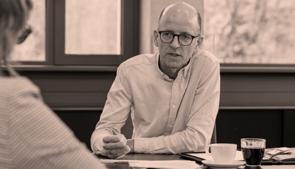 Schwarz-Wei&szlig;-Aufnahme von Thomas Melmer w&auml;hrend eines Interviews. Er sitzt an einem Tisch mit Kaffeetasse und Unterlagen und spricht konzentriert mit einer Person im Vordergrund, die nur von hinten zu sehen ist.