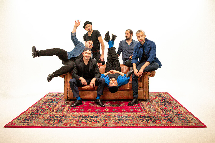 Die sechsk&ouml;pfige Band Les Yeux d'la T&ecirc;te in einer spielerischen, unkonventionellen Pose auf und um ein braunes Chesterfield-Sofa auf einem gemusterten Orientteppich vor wei&szlig;em Hintergrund