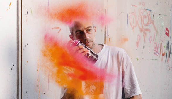 Ein Mann in einem wei&szlig;en T-Shirt spr&uuml;ht mit einer Spr&uuml;hdose leuchtend orangefarbene und rosa Farbe in die Luft. Die Farbwolke schwebt vor seinem Gesicht. Im Hintergrund ist eine helle, mit Graffiti und Farbspritzern versehene Wand zu sehen.