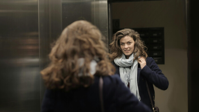 Eine Frau steht in einem Aufzug und betrachtet sich im Spiegel, w&auml;hrend sie ihren Schal richtet. Das Bild symbolisiert Arbeitsalltag und Themen wie Lohntransparenz und Teilzeitarbeit.