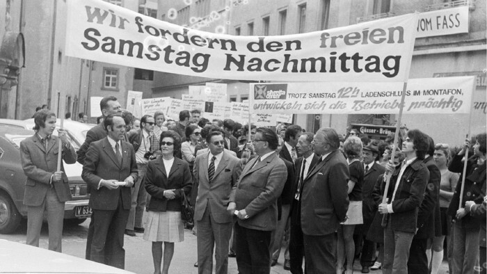 Schwarz-Wei&szlig;-Foto einer Kundgebung auf einer Stra&szlig;e. Eine Menschenmenge h&auml;lt Transparente hoch, auf dem vordersten steht: &bdquo;Wir fordern den freien Samstag Nachmittag&ldquo;. Im Hintergrund ist ein weiteres Transpartent mit Text wie &bdquo;Trotz Samstag 12h Ladenschluss im Montafon, entwickeln sich die Betriebe pr&auml;chtig&ldquo; zu sehen.
