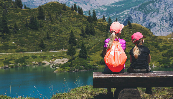 Zwei Mädchen sitzen auf einer Bank vor einem Bergsee.