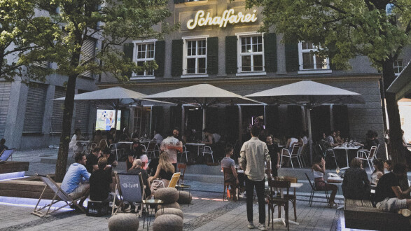 Abendliche Au&szlig;enansicht des Geb&auml;udes der Schaffarei im AK Quartier. Im geschm&uuml;ckten Au&szlig;enbereich sitzen zahlreiche Personen an Tischen unter gro&szlig;en wei&szlig;en Schirmen sowie auf Liegest&uuml;hlen. &Uuml;ber der Fassade leuchtet der Schriftzug &bdquo;Schaffarei&ldquo; in gelber Neonschrift.