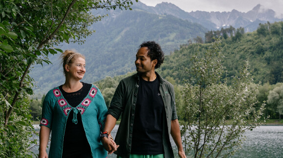 Ein l&auml;chelndes Paar h&auml;lt H&auml;ndchen und blickt sich liebevoll an, w&auml;hrend es in einer alpinen Landschaft spazieren geht. Links steht eine Frau mit hochgesteckten blonden Haaren, einem Haarband und einer markanten t&uuml;rkisfarbenen Strickjacke mit rosa-wei&szlig;en H&auml;kelmustern. Rechts steht ein Mann mit dunklen, lockigen Haaren, einem schwarzen T-Shirt und einem offenen, dunkelgr&uuml;nen Hemd. Im Hintergrund erstreckt sich ein ruhiger See, hinter dem dicht bewaldete Bergh&auml;nge und schroffe, wolkenverhangene Gebirgsz&uuml;ge aufragen. Am linken und rechten Bildrand rahmen gr&uuml;ne Str&auml;ucher und B&auml;ume die Szene ein.