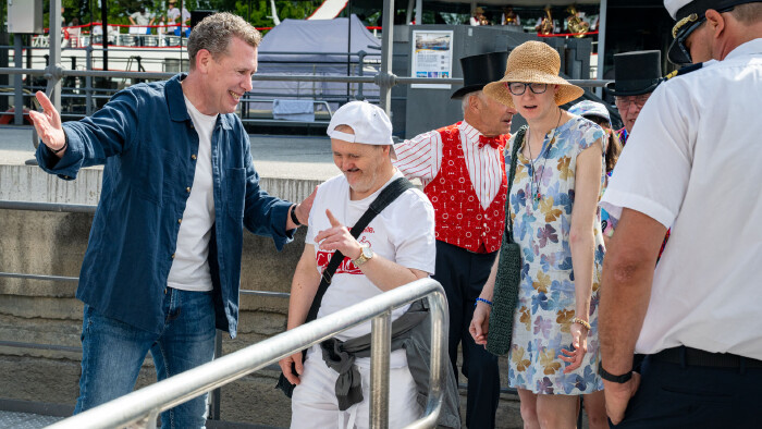 AK Pr&auml;sident Bernhard Heinzle begr&uuml;&szlig;t am Hafen mehrere G&auml;ste der j&auml;hrlichen AK Schifffahrt f&uuml;r Menschen mit Behinderung auf dem Bodensee. Er steht l&auml;chelnd neben den Teilnehmer:innen und gestikuliert einladend. Im Hintergrund sind weitere G&auml;ste, Helfer:innen und ein Schiff zu sehen. Die Szene vermittelt eine freundliche und herzliche Atmosph&auml;re.