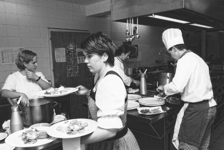 Schwarz-Wei&szlig;-Aufnahme aus den 1980er-Jahren: Gesch&auml;ftiger Betrieb in einer Restaurantk&uuml;che. Eine Servicekraft tr&auml;gt mehrere Teller mit Speisen, eine zweite steht daneben. Im Hintergrund arbeiten zwei K&ouml;che am Herd. An der Decke h&auml;ngen Sch&ouml;pfkellen, die Arbeitsfl&auml;chen sind mit T&ouml;pfen und Tellern belegt.