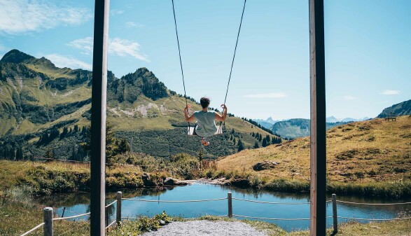 Das Foto zeigt eine Frau auf einer Schaukel. Direkt vor ihr liegt ein schöner, blauer Bergsee, im Hintergrund sieht man Berggipfel, grüne Wiesen und blauen Himmel.