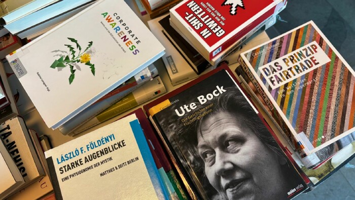  Ein Tisch mit einer vielfältigen Auswahl an Büchern, die in unterschiedlichen Sprachen und Formaten präsentiert sind. Die Bücher sind teils gestapelt, teils aufgefächert und zeigen ein breites Spektrum an Themen aus Bereichen wie Gesellschaft, Wirtschaft, Mystik und Humanität. Die Gestaltung der Buchcover reicht von schlicht bis farbenfroh und vermittelt einen Eindruck von kultureller und inhaltlicher Vielfalt.