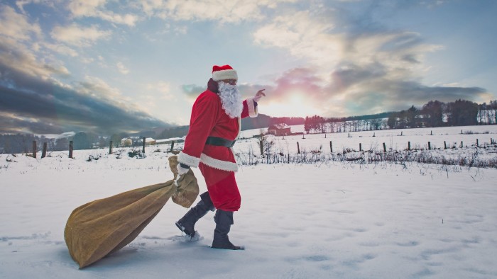 Der Weihnachtsmann mit einer Tüte voller Geschenke geht über ein schneebedecktes Feld, am Horizont ein Winterwald und Sonnenlicht.