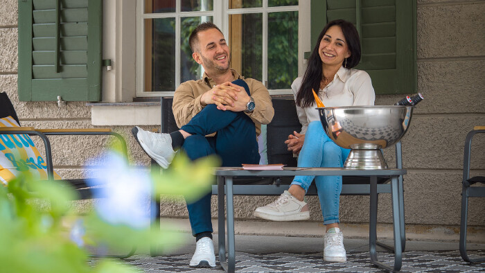 Portr&auml;t von Adrien und Alyssa, den neuen P&auml;chtern der Schaffarei Gastronomie x Aventure im AK Quartier. Beide sitzen entspannt und fr&ouml;hlich auf einer schwarzen Gartenbank im Au&szlig;enbereich vor einem Fenster mit gr&uuml;nen Fensterl&auml;den. Links sitzt Adrien im beigen Hemd, dunkler Hose und wei&szlig;en Sneakern, die Beine &uuml;bereinandergeschlagen. Rechts sitzt Alyssa mit langen, dunklen Haaren, wei&szlig;er Bluse, hellblauen Jeans und ebenfalls wei&szlig;en Sneakern. Auf dem dunklen Tisch vor ihnen steht eine gro&szlig;e, gl&auml;nzende Metallschale zur Flaschenk&uuml;hlung. Im verschwommenen Vordergrund sind gr&uuml;ne Bl&auml;tter zu sehen.