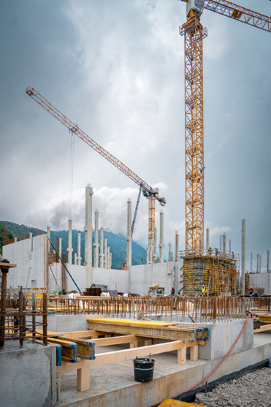 Eine Weitwinkelaufnahme einer gro&szlig;en Baustelle unter einem dicht bew&ouml;lkten Himmel. Im Vordergrund sind gegossene Betonfundamente mit freiliegenden, vertikalen Bewehrungsst&auml;hlen sowie Holzverschalungen und Bauelemente zu sehen. Dahinter erstrecken sich hohe, im Bau befindliche Betonw&auml;nde und zahlreiche aufragende Betons&auml;ulen. Die Baustelle wird von zwei gro&szlig;en Turmdrehkranen dominiert: Ein m&auml;chtiger, gelber Kran steht auf der rechten Seite, w&auml;hrend ein zweiter, orange-roter Kran diagonal durch die obere linke Bildh&auml;lfte ragt. Im Hintergrund sind bewaldete Berge im Dunst zu erkennen.