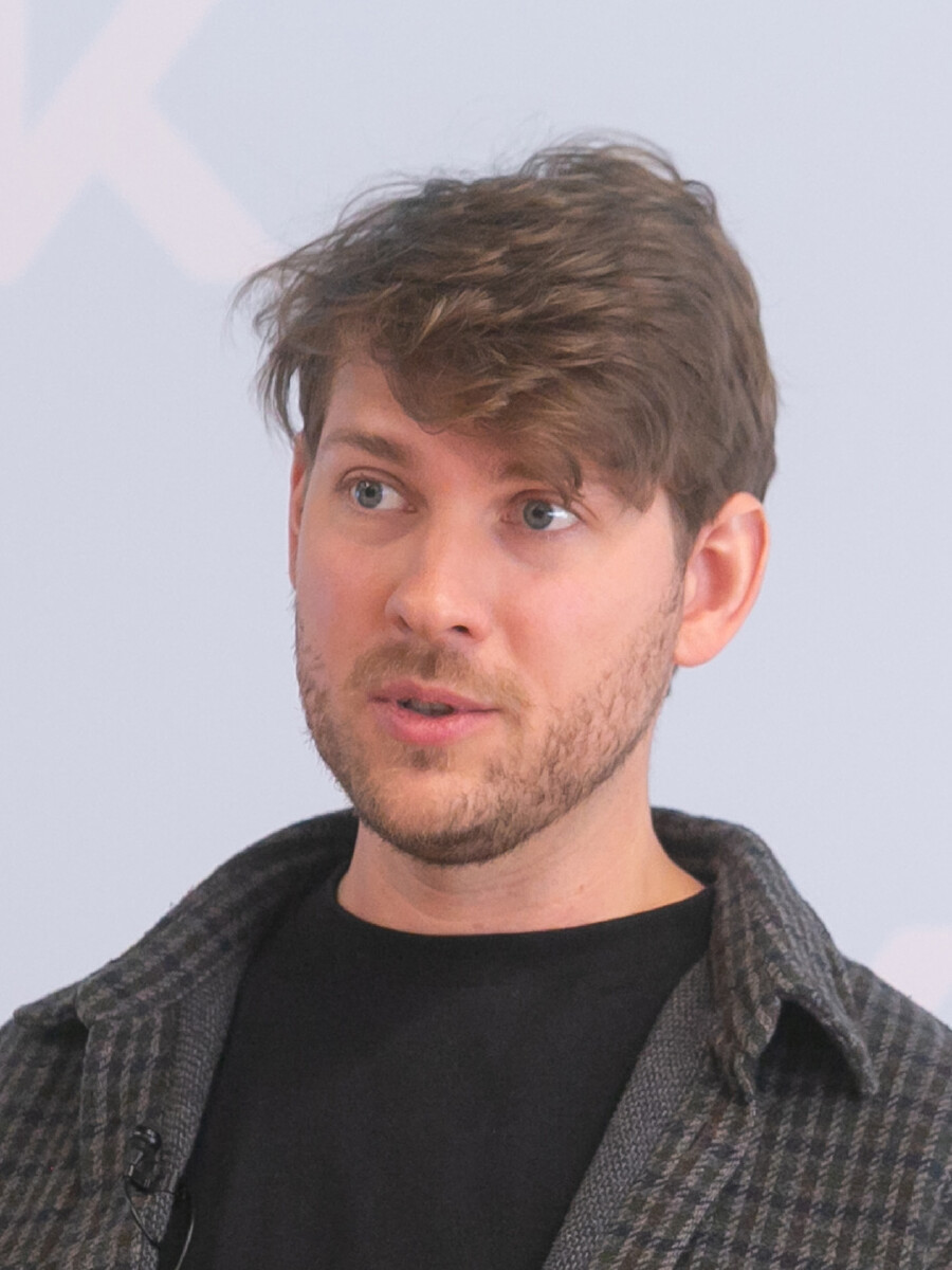 Portr&auml;tfoto von Dominic G&ouml;tz, &Ouml;konom der Arbeiterkammer Vorarlberg. Er spricht in die Kamera, tr&auml;gt ein schwarzes T-Shirt unter einem grauen Karojackett und hat kurzes, braunes Haar sowie einen kurzen Bart. Der Hintergrund ist hell und schlicht. Foto: Dietmar Mathis.