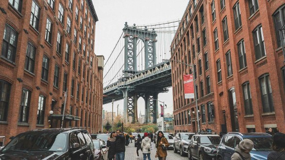 Mehrere Menschen stehen auf einer Straße in New York, im Hintergrund sieht man die berühmte Brooklyn Bridge.