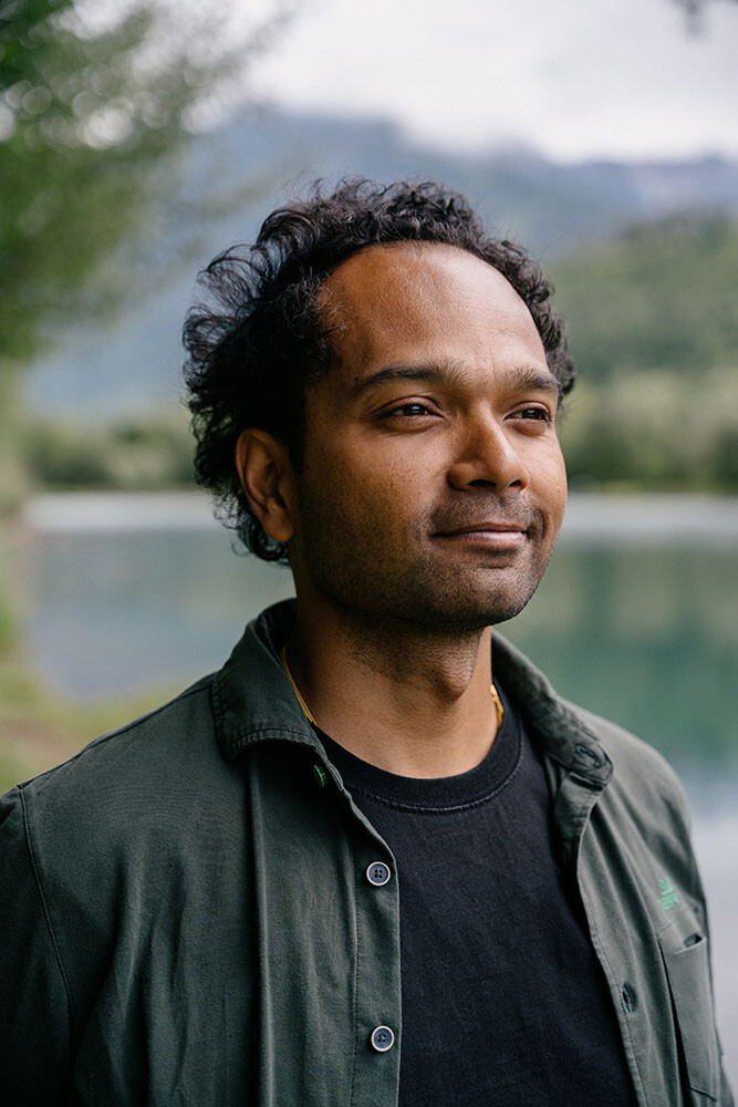Ein Portr&auml;t von Ankur Jaiswal im Halbprofil vor einer Naturkulisse. Er blickt mit einem sanften, zufriedenen L&auml;cheln nach rechts am Kamerachor vorbei in die Ferne. Er hat eine dunklere Hautfarbe, dunkles, dichtes Lockenhaar und tr&auml;gt ein schwarzes T-Shirt unter einem offenen, dunkelgr&uuml;nen Hemd. Der Hintergrund zeigt einen leicht verschwommenen See und gr&uuml;ne, bewaldete Berge unter einem bew&ouml;lkten Himmel.