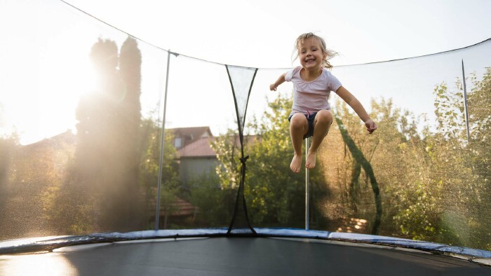 Ein lachendes Mädchen springt bei Sonnenschein auf einem Gartentrampolin mit Sicherheitsnetz – im Hintergrund sind Bäume und Hausdächer zu sehen.