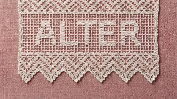 Querformatiger Detailausschnitt der H&auml;kelspitze mit dem Schriftzug &bdquo;ALTER&ldquo; auf altrosa Textilgrund. Die untere Zackenkante der Spitze ist vollst&auml;ndig sichtbar.