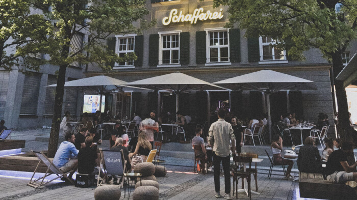 Abendliche Au&szlig;enansicht des Geb&auml;udes der Schaffarei im AK Quartier. Im geschm&uuml;ckten Au&szlig;enbereich sitzen zahlreiche Personen an Tischen unter gro&szlig;en wei&szlig;en Schirmen sowie auf Liegest&uuml;hlen. &Uuml;ber der Fassade leuchtet der Schriftzug &bdquo;Schaffarei&ldquo; in gelber Neonschrift.
