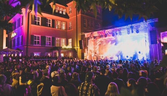 Ein große Menschenmenge steht vor einer beleuchteten Bühne, auf der eine Band gerade ein Konzert gibt.