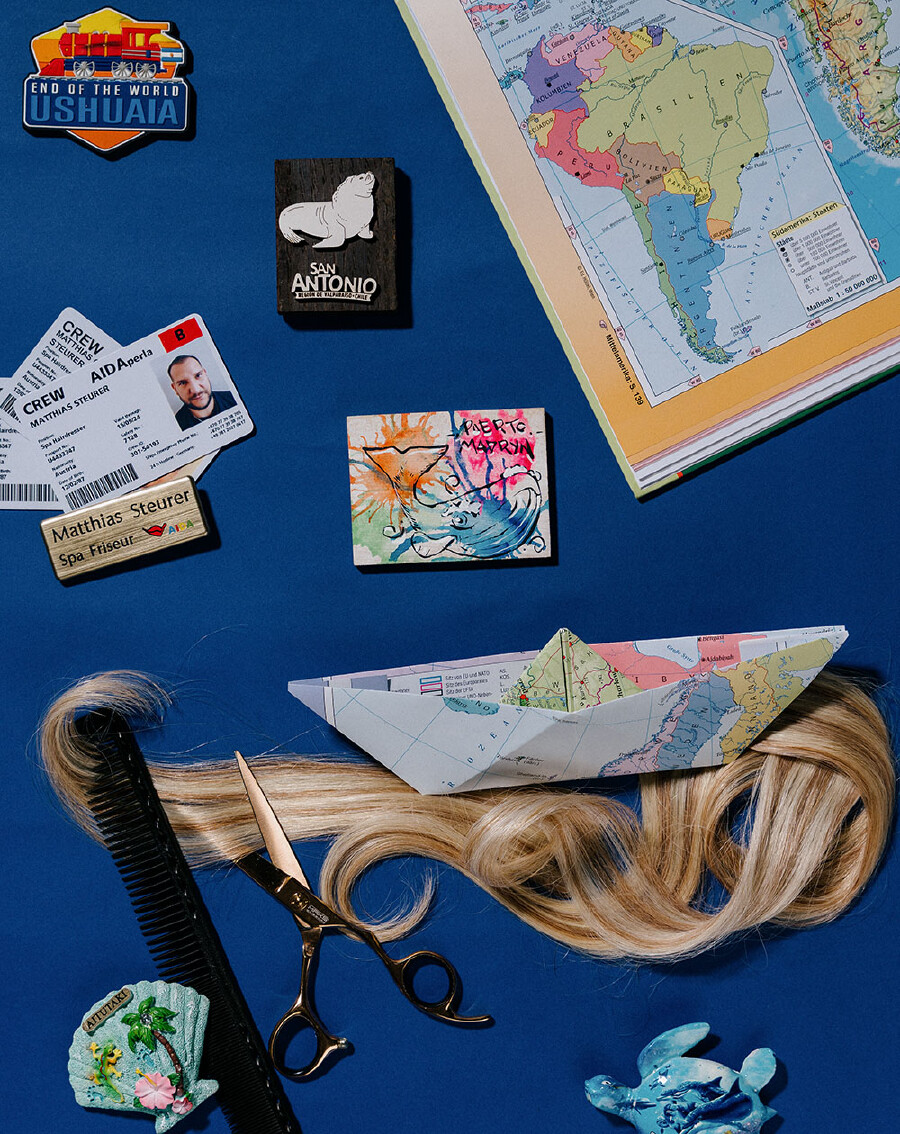 Eine kunstvolle Draufsicht vor einem kr&auml;ftig blauen Hintergrund, auf dem verschiedene Souvenirs von Seereisen und Utensilien des Friseurhandwerks arrangiert sind.  Im oberen Bereich sieht man eine ge&ouml;ffnete Buchseite mit einer farbigen Landkarte von S&uuml;damerika sowie diverse K&uuml;hlschrankmagnete, darunter einer mit einer roten Lokomotive (&bdquo;Ushuaia&ldquo;), einer mit einem Seel&ouml;wen (&bdquo;San Antonio&ldquo;) und einer mit einer bunten Wal-Illustration (&bdquo;Puerto Madryn&ldquo;). Links liegen eine wei&szlig;e Crew-ID-Karte des Kreuzfahrtschiffs &bdquo;AIDAperla&ldquo; mit dem Namen Matthias Steurer und einem Portr&auml;tfoto sowie sein goldenes Namensschild mit der Aufschrift &bdquo;Matthias Steurer &ndash; Spa Friseur&ldquo;.  Im unteren Drittel zieht sich eine lange, geschwungene Str&auml;hne aus blondem Echthaar quer durch das Bild. Auf den Haaren sitzt ein aus einer Landkarte gefaltetes Papierschiffchen. Ganz unten links liegen eine goldene Friseurschere, ein schwarzer Kamm und ein kleiner Muschel-Magnet, w&auml;hrend unten rechts eine kleine, blau gemusterte Schildkr&ouml;tenfigur platziert ist.