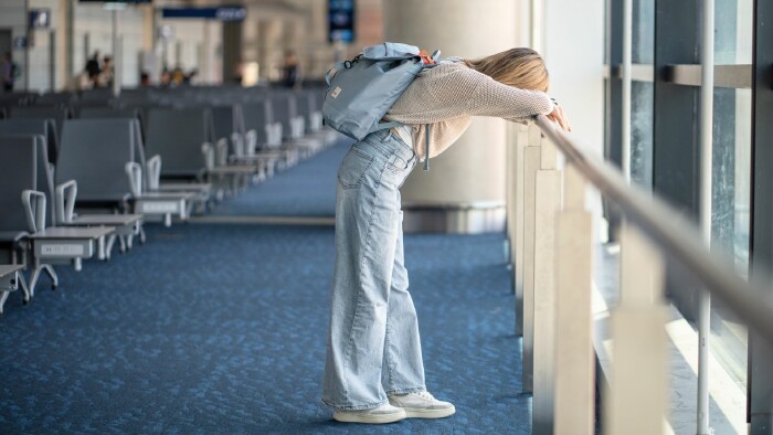 Eine junge Frau mit Rucksack lehnt entt&auml;uscht und ersch&ouml;pft am Gel&auml;nder einer leeren Flughafen-Wartehalle und st&uuml;tzt ihren Kopf auf die Arme.