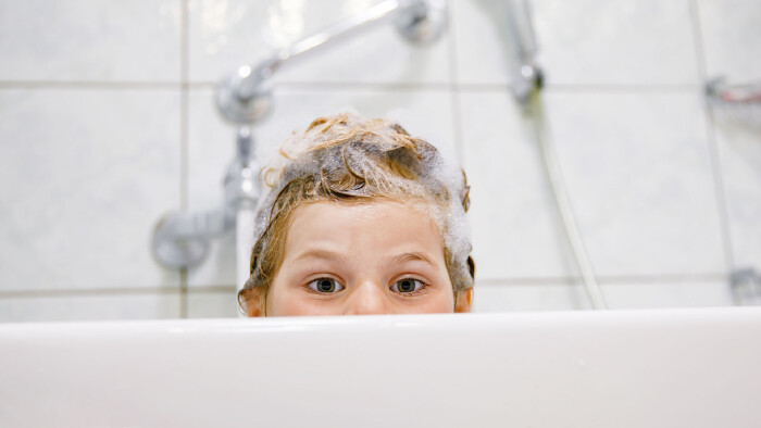 Ein Kind blickt mit nassen Haaren voller Shampoo-Schaum über den Rand einer Badewanne in die Kamera.