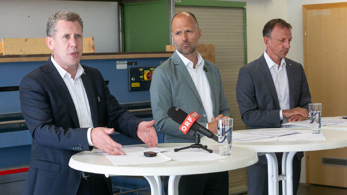 Eine Aufnahme von einer Pressekonferenz in einer Werkstattumgebung. Drei M&auml;nner in Anz&uuml;gen stehen nebeneinander hinter zwei wei&szlig;en Stehtischen. Der Mann ganz links im dunkelblauen Anzug spricht engagiert mit ge&ouml;ffneten H&auml;nden; vor ihm liegen Dokumente auf dem Tisch. Der mittlere Mann in einem graugr&uuml;nen Sakko und wei&szlig;em Hemd schaut aufmerksam zu ihm her&uuml;ber. Auf seinem Tisch steht ein Tischmikrofon mit dem roten ORF-Logo sowie ein Glas Wasser. Der dritte Mann ganz rechts im dunkelgrauen Anzug blickt mit ernstem Ausdruck nach unten auf seine Unterlagen. Im Hintergrund sind industrielle Werkstattm&ouml;bel, darunter eine blaue Maschine und ein gr&uuml;ner Schrank, zu sehen.