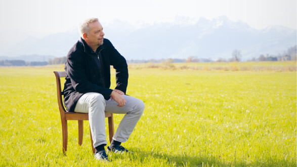 Oliver Loacker sitzt auf einem Stuhl auf einer Wiese, im Hintergrund ein Bergpanorama.