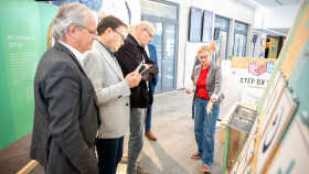 Eine Mit-Organisatorin einer Demokratie-Ausstellung erkl&auml;rt mehreren Besuchern eine interaktive Ausstellungsstation. Die Frau steht rechts und zeigt auf ein Exponat, w&auml;hrend drei M&auml;nner aufmerksam zuh&ouml;ren und Infomaterial in den H&auml;nden halten. Die Szene spielt in einem hellen Ausstellungsraum mit Infotafeln zum Thema Demokratie.