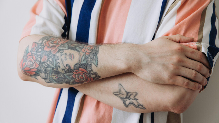 Nahaufnahme verschr&auml;nkter Arme einer Person im gestreiften Hemd. Der linke Unterarm ist gro&szlig;fl&auml;chig t&auml;towiert mit Rosen, einem Frauengesicht und Schriftz&uuml;gen im traditionellen Tattoo-Stil. Auf dem rechten Unterarm ist ein kleineres Stern-Tattoo zu sehen.