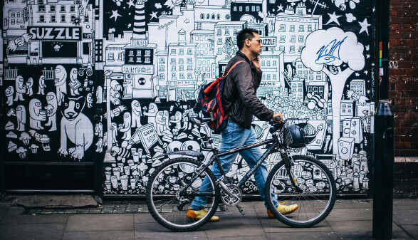 Eine Person mit Rucksack schiebt ein Fahrrad auf einem Gehweg an einer gro&szlig;en schwarz-wei&szlig; bemalten Wand mit vielen Comic-artigen Figuren und Stadtmotiven vorbei.