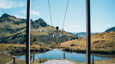 Das Foto zeigt eine Frau auf einer Schaukel. Direkt vor ihr liegt ein schöner, blauer Bergsee, im Hintergrund sieht man Berggipfel, grüne Wiesen und blauen Himmel.