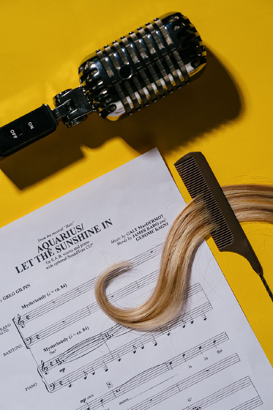 Eine Draufsicht vor einem leuchtend gelben Hintergrund, auf der Symbole f&uuml;r Musik und das Friseurhandwerk kombiniert sind.  Im oberen Bereich liegt horizontal ein gl&auml;nzendes, silbernes Vintage-Mikrofon mit einem schwarzen Ein- und Ausschalter. Darunter befindet sich ein wei&szlig;es Notenblatt mit den Noten und dem Titel des Songs &bdquo;Aquarius / Let the Sunshine In&ldquo; aus dem Musical &bdquo;Hair&ldquo;. Quer &uuml;ber das Notenblatt schmiegt sich eine lange, sanft geschwungene blonde Haarstr&auml;hne. Ein dunkelbrauner Stielkamm steckt im rechten Teil der Str&auml;hne, als ob das Haar gerade gek&auml;mmt w&uuml;rde.