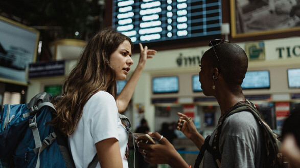 Zwei junge Frauen mit Reiserucks&auml;cken stehen in einem Bahnhof oder Flughafen und sprechen miteinander. Eine h&auml;lt ein Smartphone in der Hand, die andere gestikuliert erkl&auml;rend. Im Hintergrund ist eine gro&szlig;e Abflugtafel sowie ein Ticketschalter zu sehen.