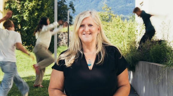 Lächelnde Sozialbetreuerin mit türkisfarbener Halskette vor grüner Bergkulisse – engagiert im Beruf und ehrenamtlich als Gruppenleiterin bei den Pfadfindern aktiv, Symbol für soziale Verantwortung und Teamgeist.